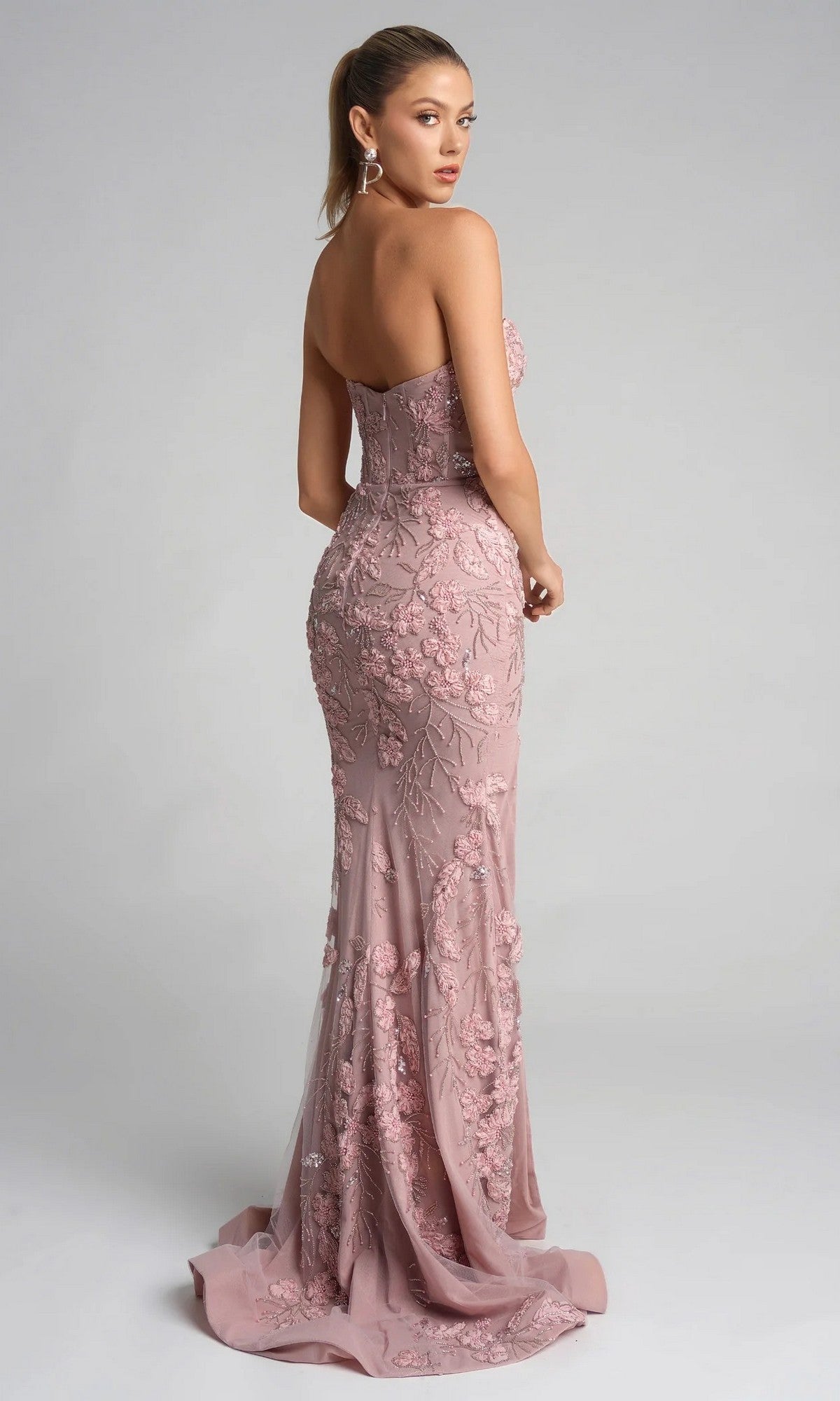 Woman wearing the Portia and Scarlett PS26074, a floor-length dusty pink gown with a strapless sweetheart neckline and floral lace appliqué, stands against a plain background looking over her shoulder.